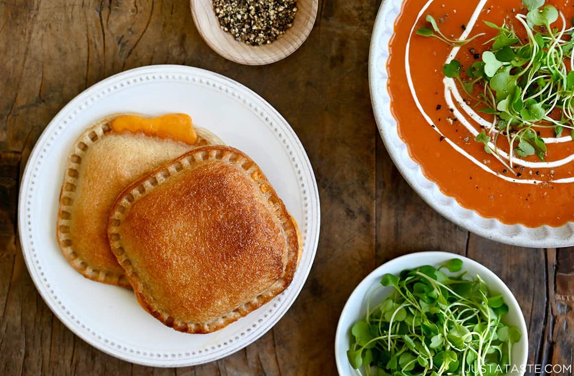 A white plate with two grilled cheese sandwiches with a bowl of tomato soup next to it