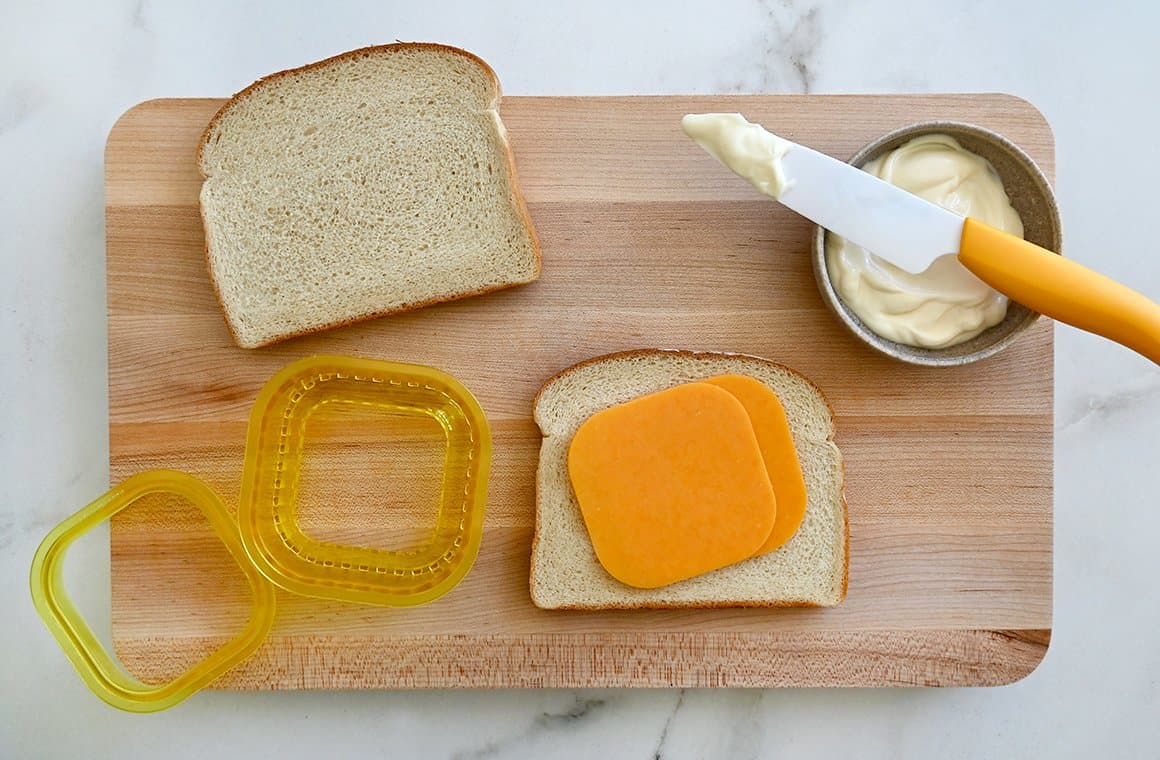A wood cutting board topped with white bread, cheddar cheese and a bowl of mayo