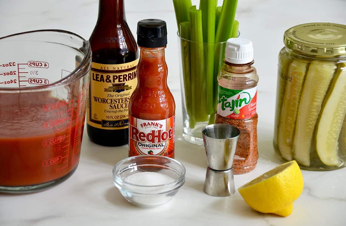 Tomato juice in a liquid measuring cup next to Worcestershire sauce, Red Hot sauce, Tajin seasoning, celery sticks, pickles in a jar, half a lemon and sugar in a small bowl