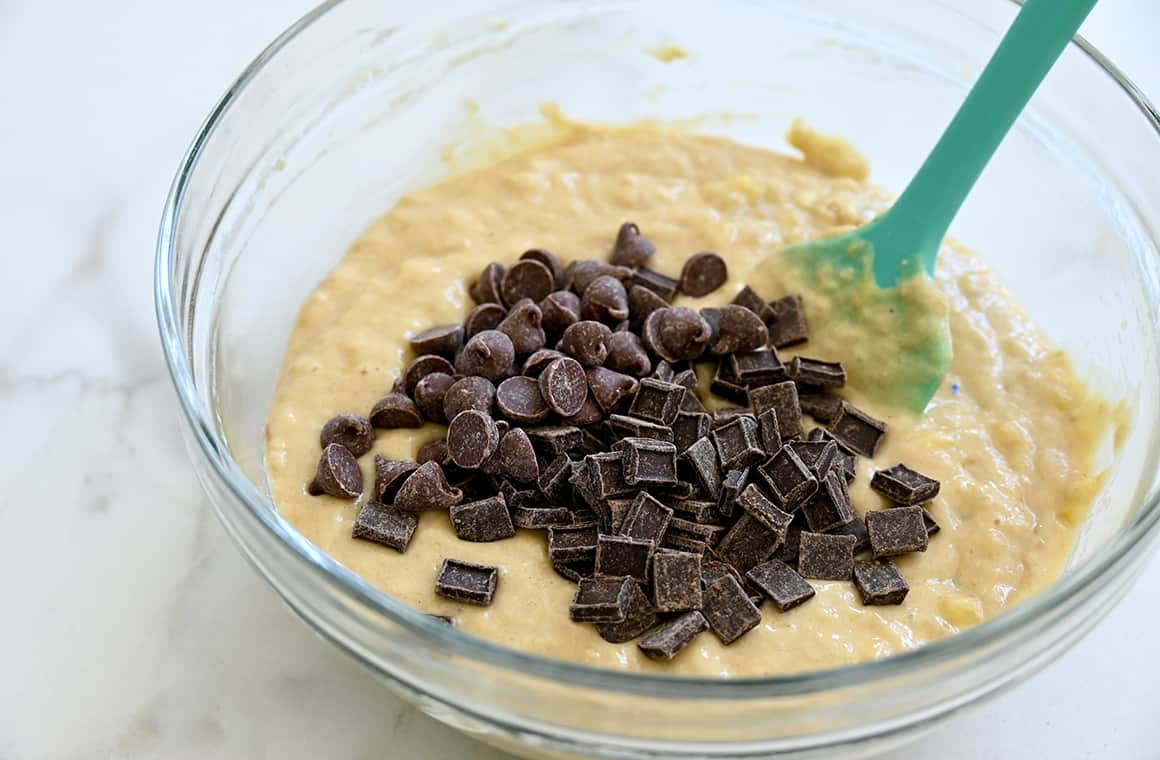 A teal spatula rests in a clear bowl containing batter, chocolate chips and chocolate chunks