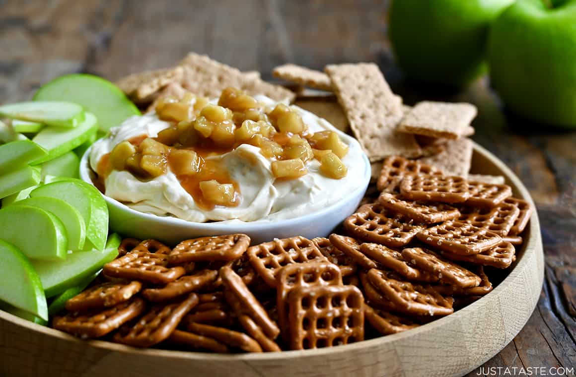 A bowl containing easy Caramel Apple Cheesecake Dip on a plate surrounded by mini pretzels, Granny Smith apple slices and graham crackers