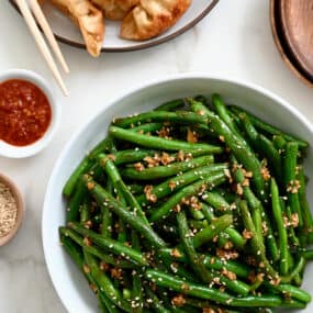 A white bowl containing Chinese Garlic Green Beans next to a plate with potstickers and chopsticks
