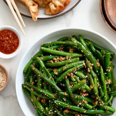 A white bowl containing Chinese Garlic Green Beans next to a plate with potstickers and chopsticks