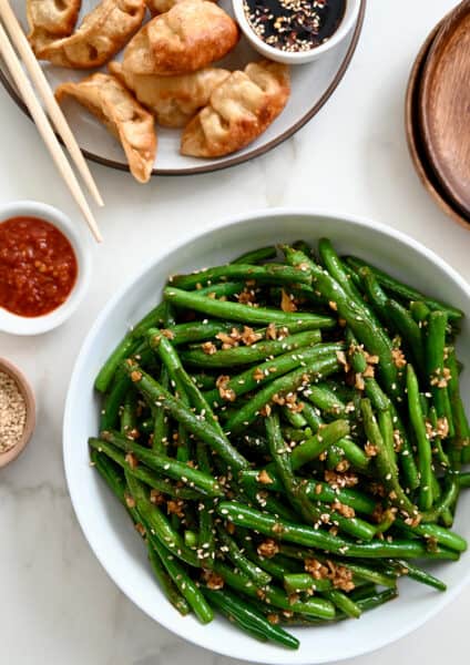 A white bowl containing Chinese Garlic Green Beans next to a plate with potstickers and chopsticks