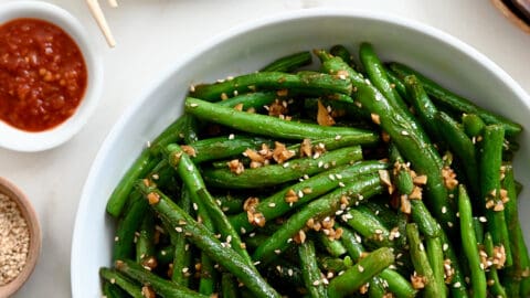 A white bowl containing Chinese Garlic Green Beans next to a plate with potstickers and chopsticks