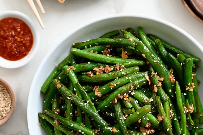 A white bowl containing Chinese Garlic Green Beans next to a plate with potstickers and chopsticks