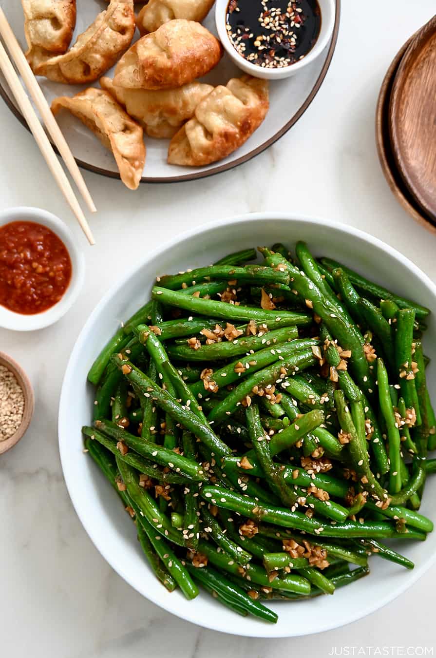 A white bowl containing Chinese Garlic Green Beans next to a plate with potstickers and chopsticks