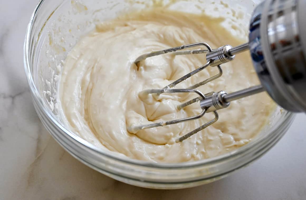 A hand mixer rests on the edge of a clear bowl that contains cream cheese filling