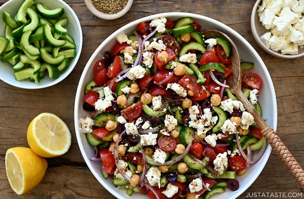 Cucumber, Tomato and Chickpea Salad with black olives and feta cheese in a white serving bowl with a wooden spoon