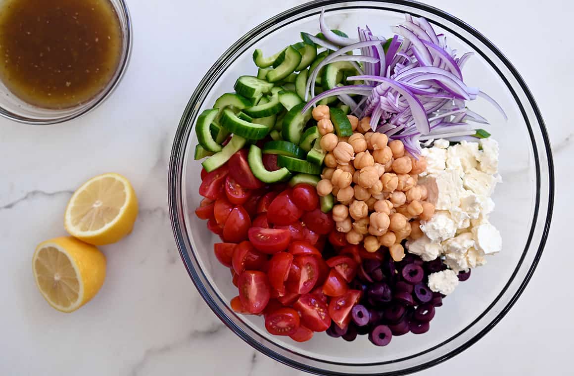 A clear bowl containing chickpeas, feta cheese, black olives, sliced red onions, halved cherry tomatoes and sliced cucumbers