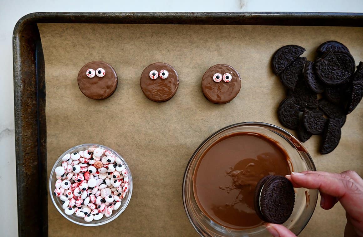 Chocolate-dipped Oreos on a baking sheet next to a bowl containing melted chocolate 