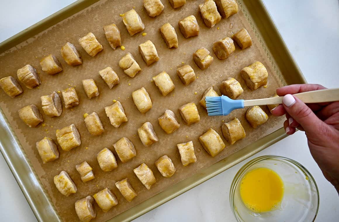 A top-down view of unbaked pretzel bites on a parchment paper-lined baking sheet being brushed with egg wash