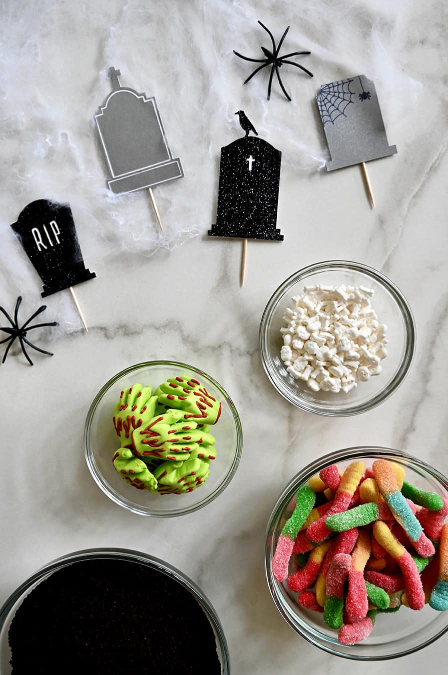 A top-down view of tombstone cake toppers next to small bowls containing candy bones, gummy worms, bloody-green gummy hands and crushed Oreos