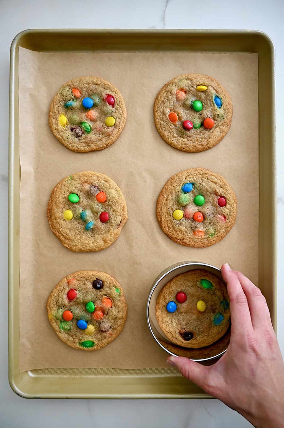 Using a round cookie cutter to swirl around a fresh-out-of-the-oven cookie on a baking sheet to make it perfectly round.
