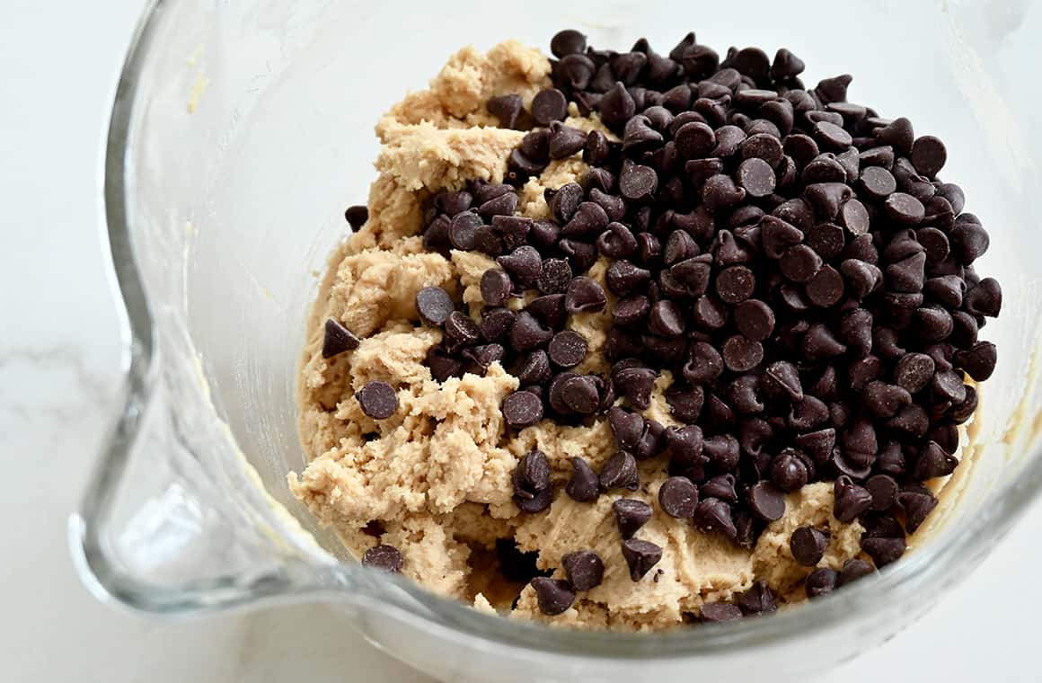 A glass mixing bowl containing cookie dough and a pile of chocolate chips