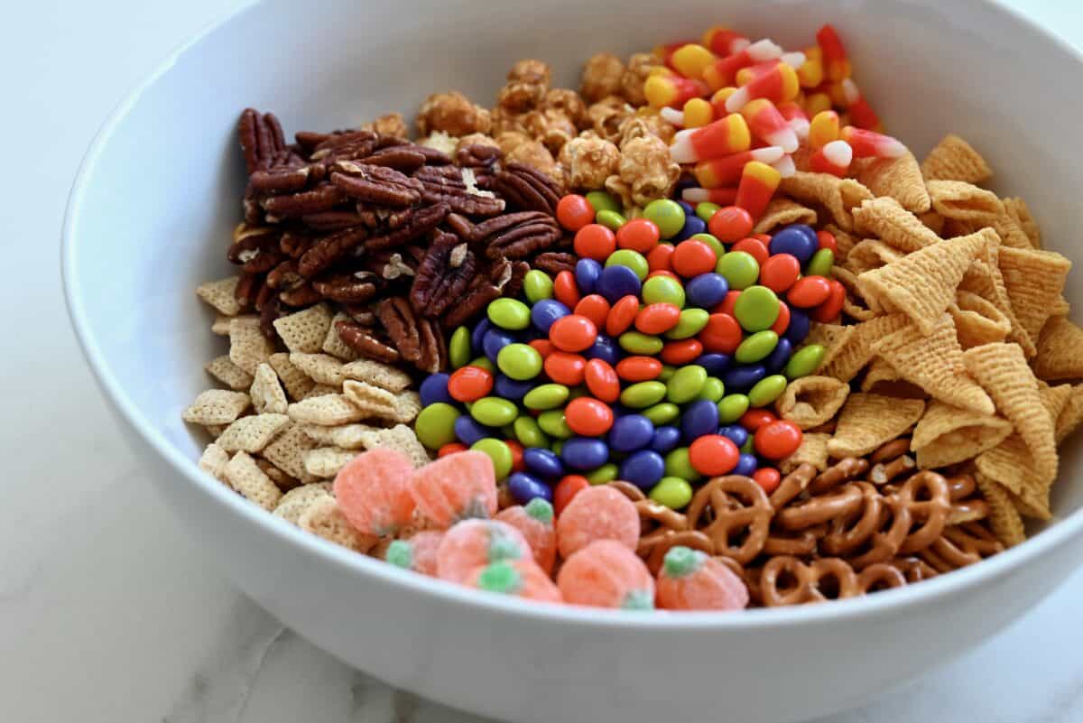 A white serving bowl containing pecans, caramel corn, candy corn, Bugles, pretzels, pumpkin candies, Chex cereal and M&M's