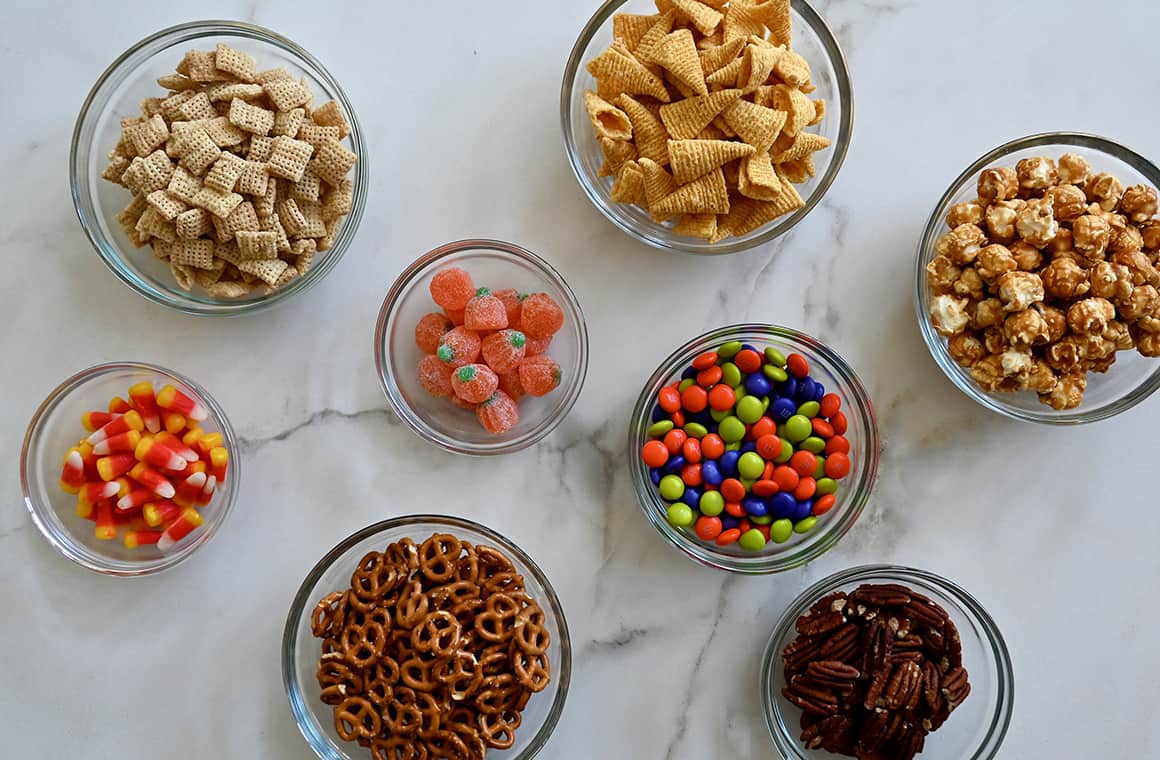 A top-down view of various sizes of small bowls containing candy corn, Chex cereal, Bugles chips, caramel corn, M&Ms, pecans and pretzels