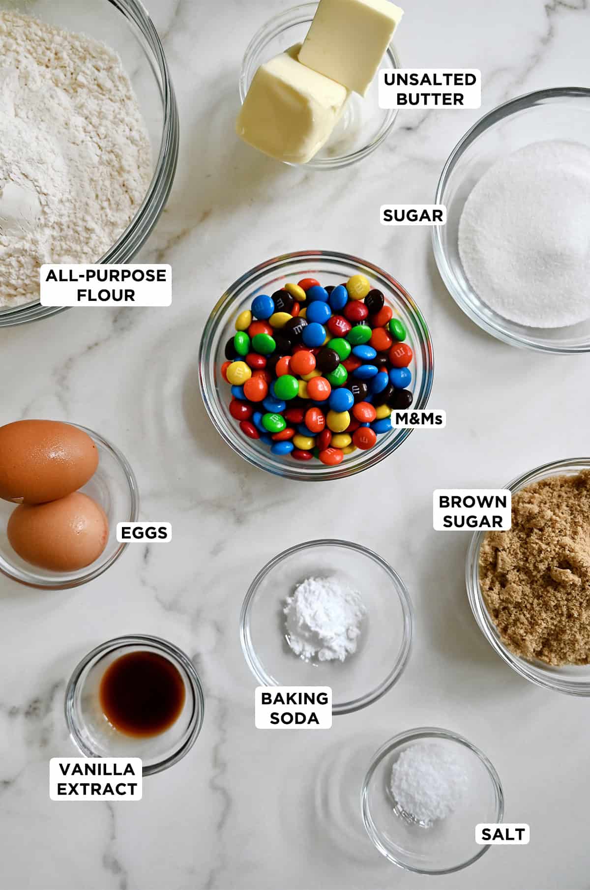 Ingredients needed to make M&M cookies in various sizes of glass bowls.