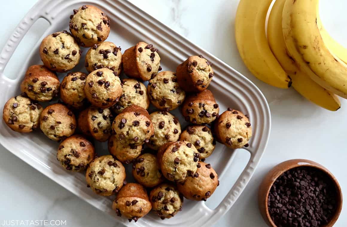 Mini Banana Chocolate Chip Muffins on a large plate next to a bowl filled with mini chocolate chips and a bunch of ripe bananas