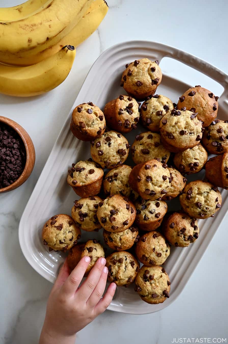 A child's hand reaches for a muffin on a platter piled high with Mini Banana Chocolate Chip Muffins