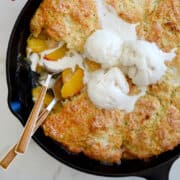 A top-down view of a cast-iron skillet containing peach cobbler topped with buttermilk biscuits and three scoops of vanilla ice cream.