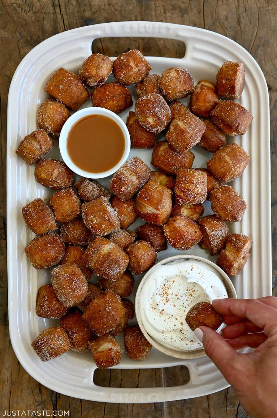 A top-down view of homemade Pumpkin Spice Pretzel Bites on a white serving platter with a hand dipping one into a bowl filled with homemade whipped cream