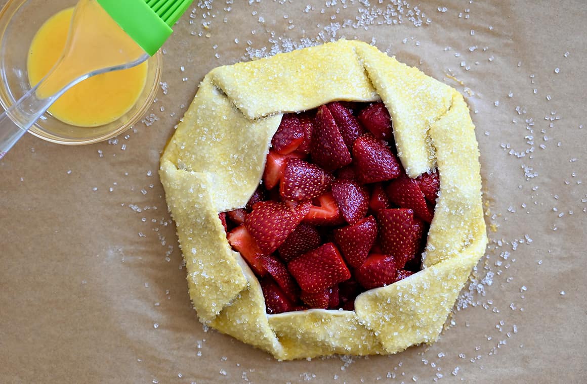 A small bowl containing egg wash next to an unbaked galette sprinkled with sanding sugar