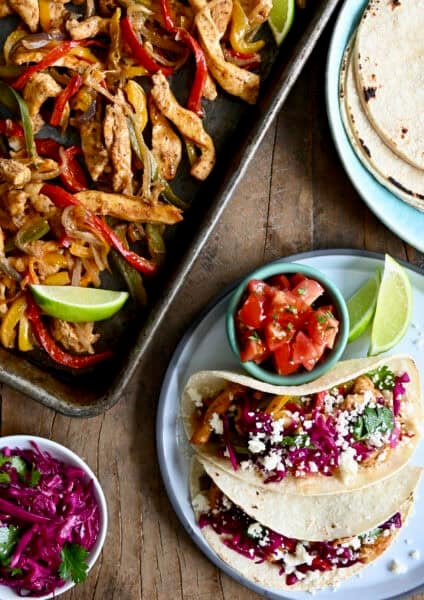 Sheet pan chicken fajitas next to a plate with tortillas and another with tacos and a small bowl containing pico de gallo.