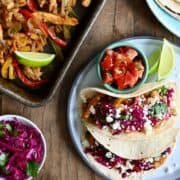 A top-down view of oven roasted Chicken Fajitas next to a plate with tacos and a plate stacked with corn tortillas.