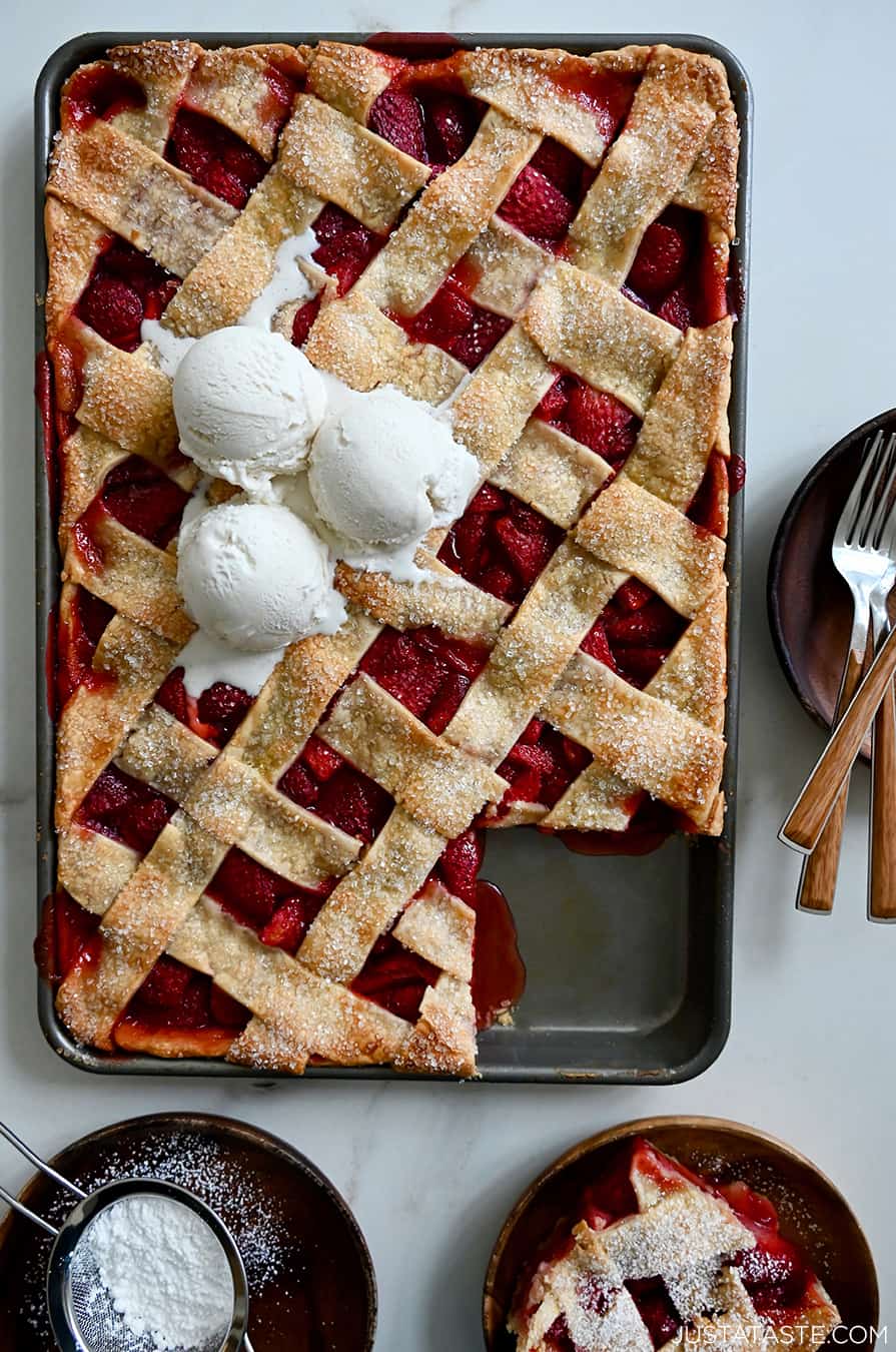 A top-down view of a Simple Strawberry Slab Pie topped with three scoops of vanilla bean ice cream and a corner piece cut out and on a plate next to it