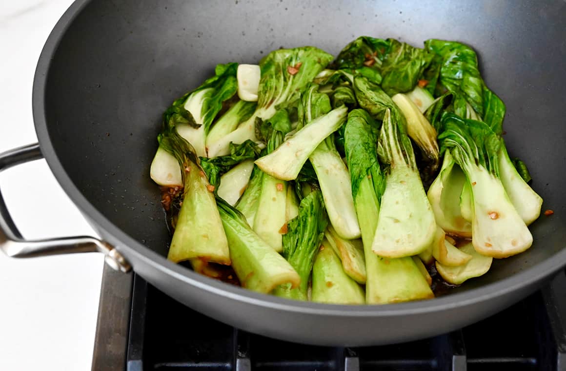 A wok on the stovetop containing bok choy leaves tossed in garlic sauce