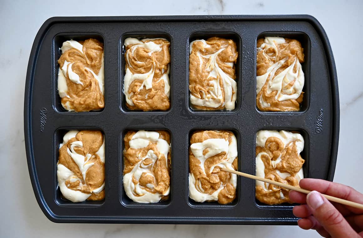 A hand uses a long skewer to swirl cheesecake into quickbread batter 