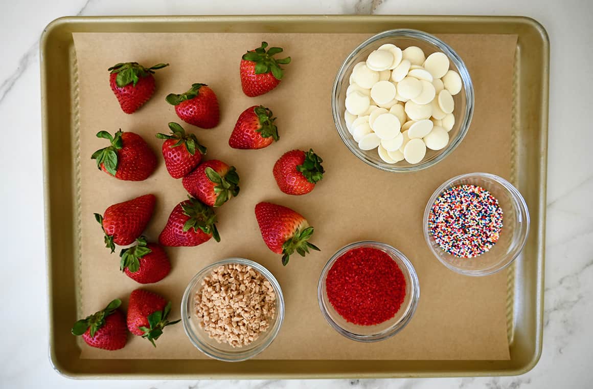 A top-down view of fresh strawberries on a baking sheet along with small bowls containing white chocolate and various sprinkles