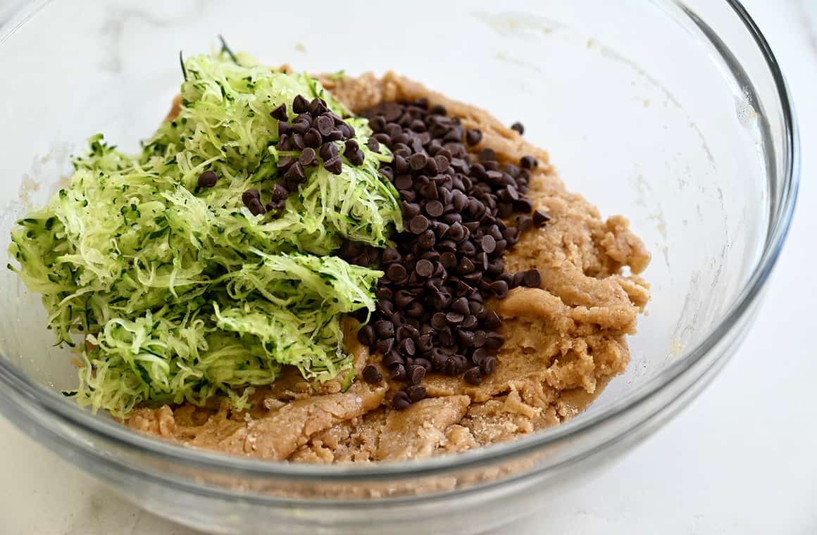 A clear bowl containing chocolate chips, batter and shredded squash