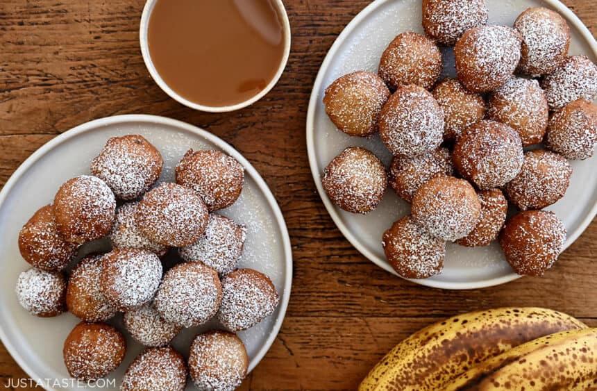 Two plates piled high with easy banana fritters dusted with powdered sugar next to a small bowl filled with caramel sauce