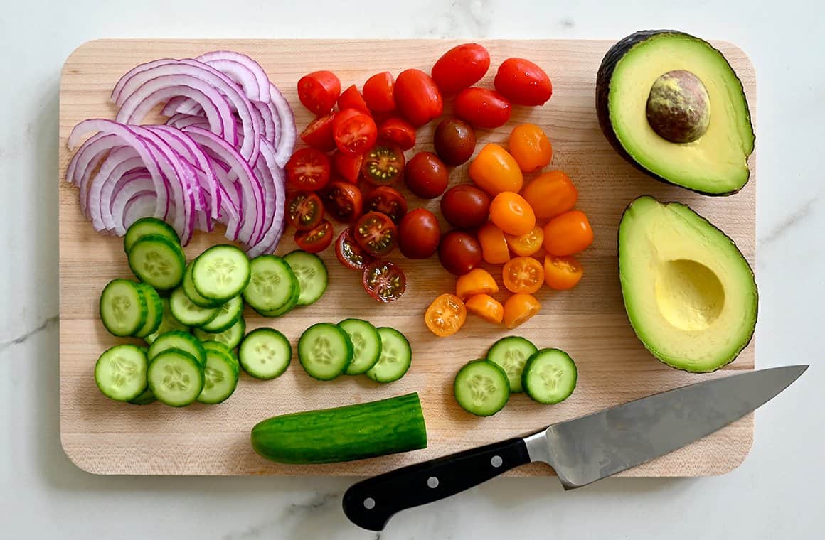 A wood cutting board with cut up vegetables, including onions, tomatoes, cucumbers and avocado
