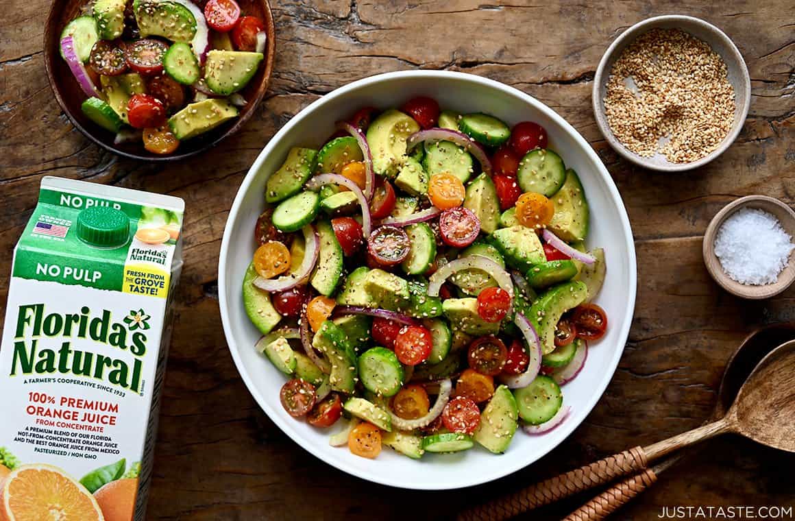 A top down view of a white bowl containing avocado salad with small bowls next to it and a carton of orange juice