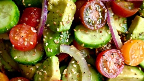 A close up view of diced avocado, red onion, cherry tomatoes and cucumbers topped with sesame seeds
