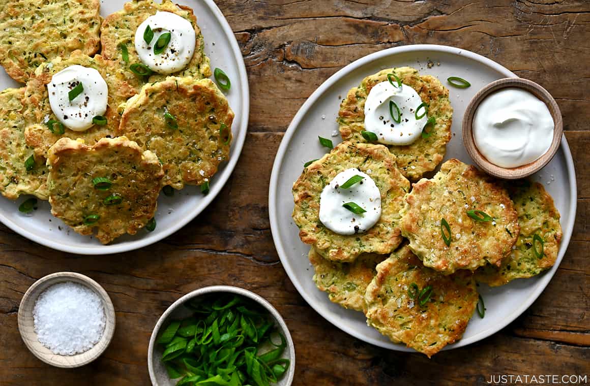 Two plates piled high with corn and zucchini fritters topped with sour cream and sliced scallions