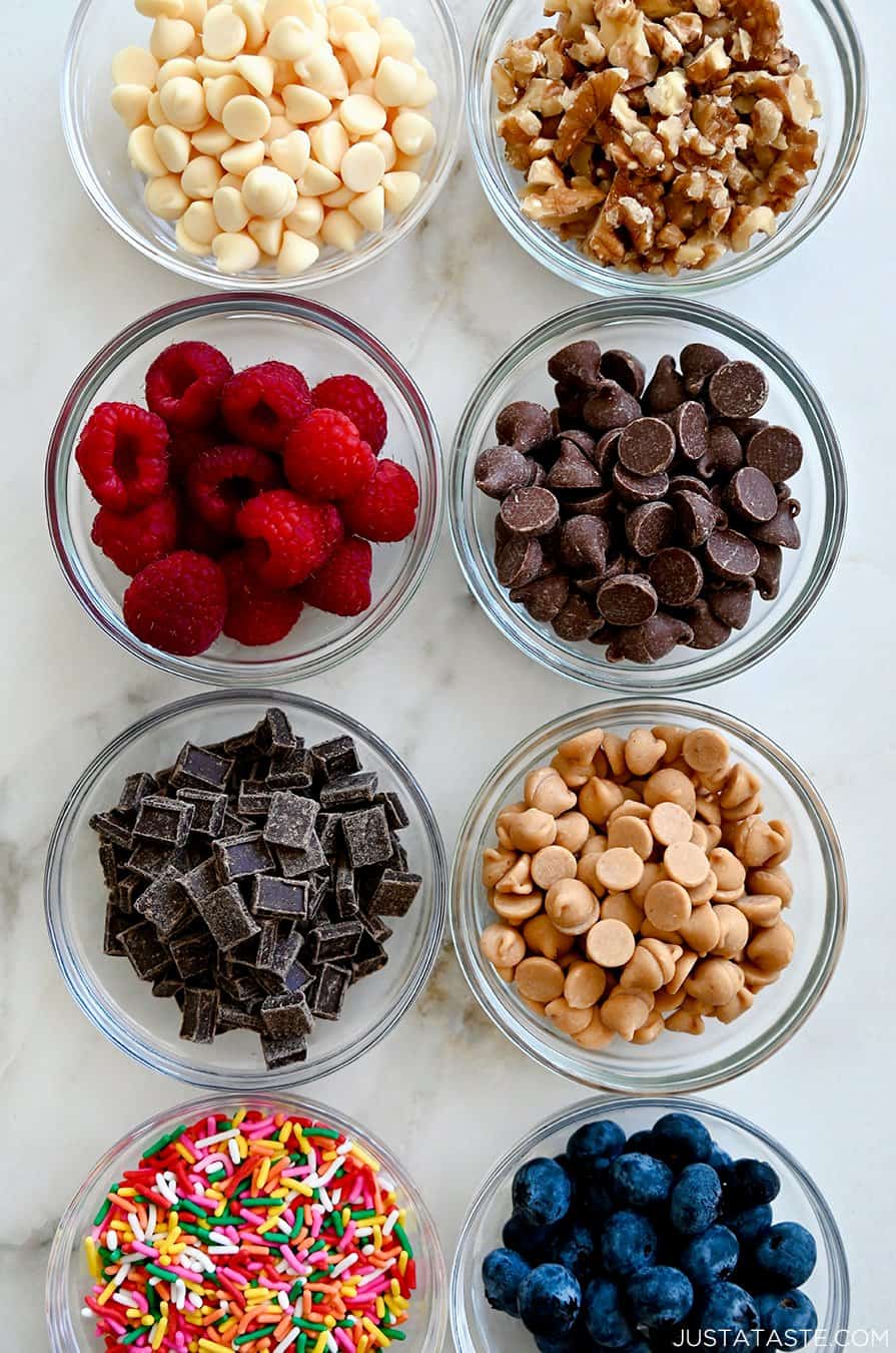 A top-down view of small bowls containing white chocolate chips, walnut pieces, raspberries, chocolate chips, chocolate chunks, peanut butter chips, rainbow sprinkles and blueberries
