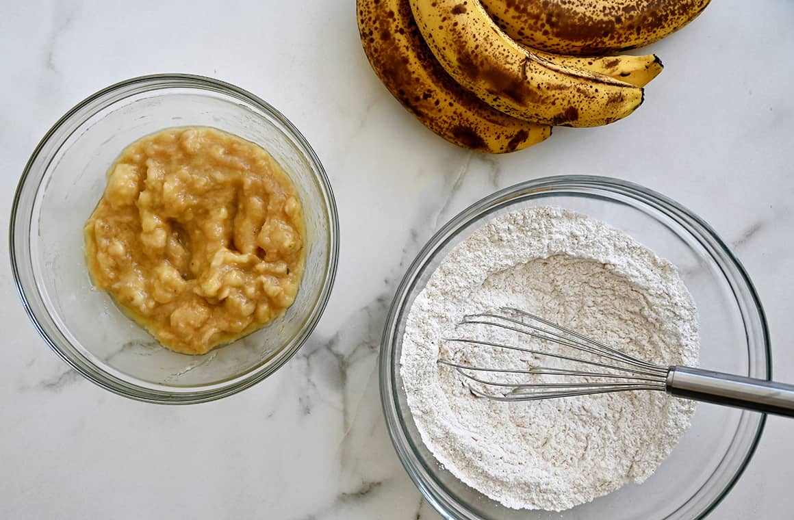 A top-down view of a bunch of bananas next to a bowl containing flour and a bowl containing mashed bananas