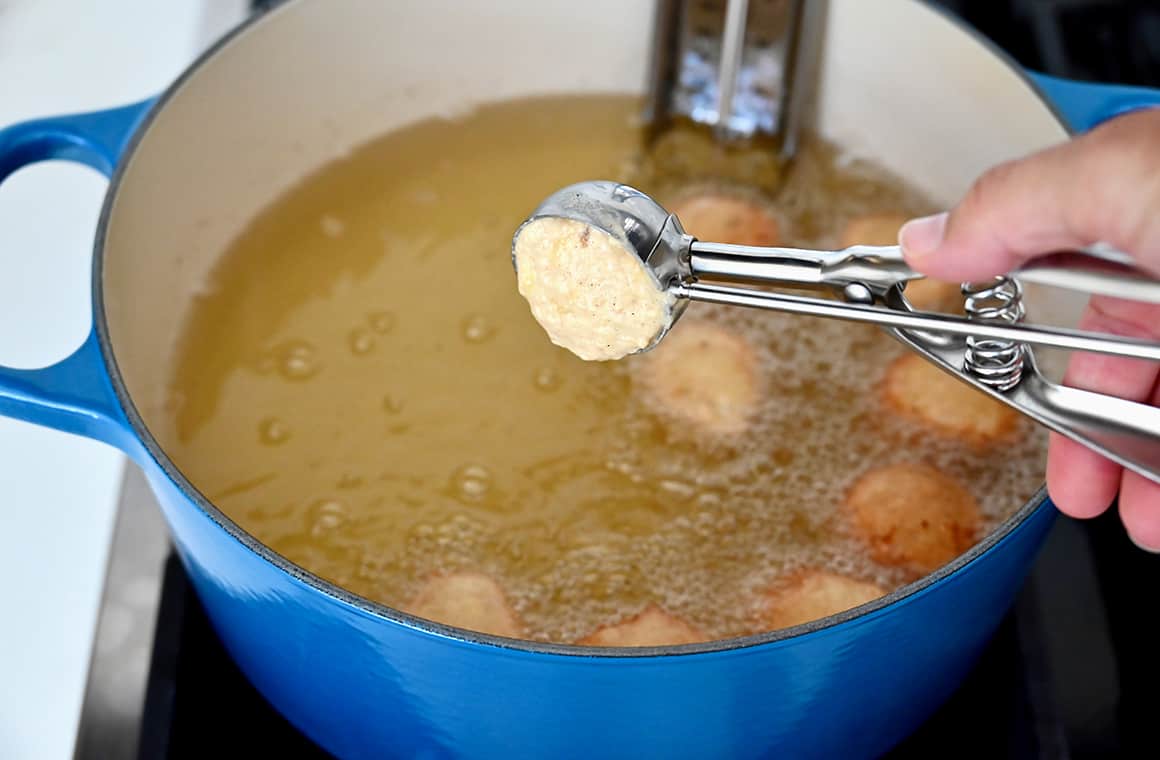 A hand holds a small ice cream scoop with batter over a stockpot containing bubbling oil