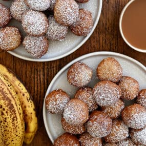 A top-down view of two plates piled high with banana fritters dusted with powdered sugar