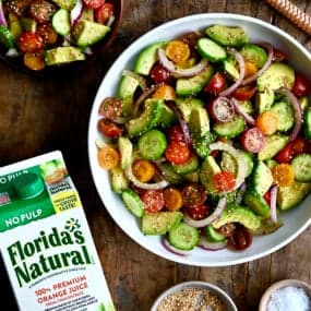 A top-down view of the best avocado and tomato salad with sliced red onions in a white bowl next to a carton of Florida's Natural orange juice