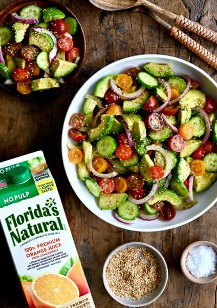 A top-down view of the best avocado and tomato salad with sliced red onions in a white bowl next to a carton of Florida's Natural orange juice