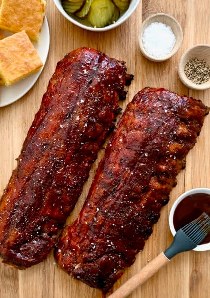 Oven-baked ribs brushed with barbecue sauce next to a plate with cornbread and a bowl containing sliced pickles.
