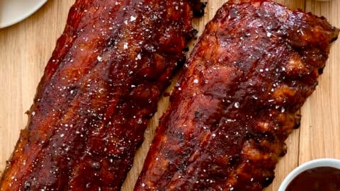 Oven-baked ribs brushed with barbecue sauce next to a plate with cornbread and a bowl containing sliced pickles.