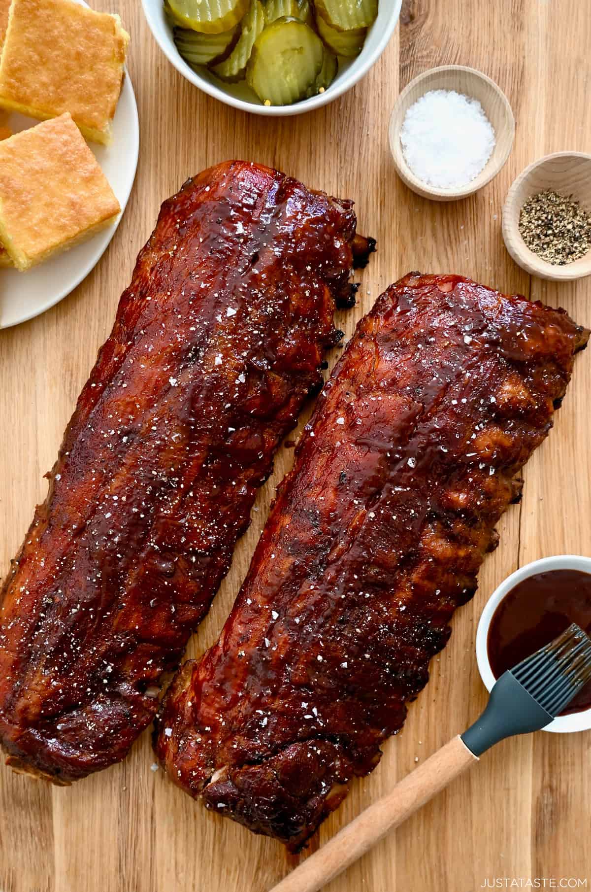 Oven-baked ribs brushed with barbecue sauce next to a plate with cornbread and a bowl containing sliced pickles.