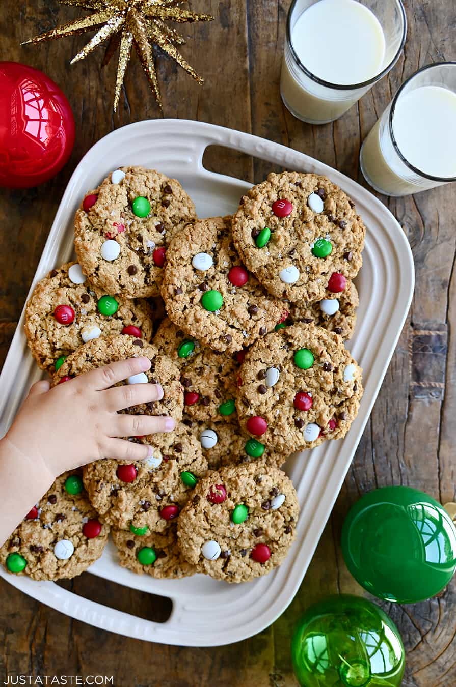 A child's hand reaches for a chewy holiday monster cookie on a platter full of cookies