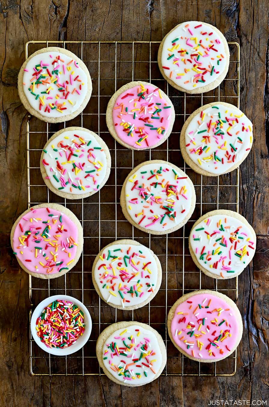 A top-down view of sugar cookies with white and pink icing and rainbow sprinkles atop a wire rack